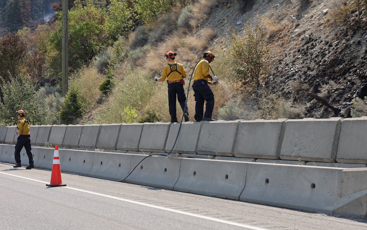 default-292 Firefighters wearing yellow are seen on the edge of a highway, with blackened ground. They are using hoses.